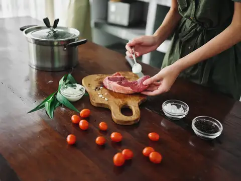 Женщина солит кусок мяса на деревянной доске среди помидоров