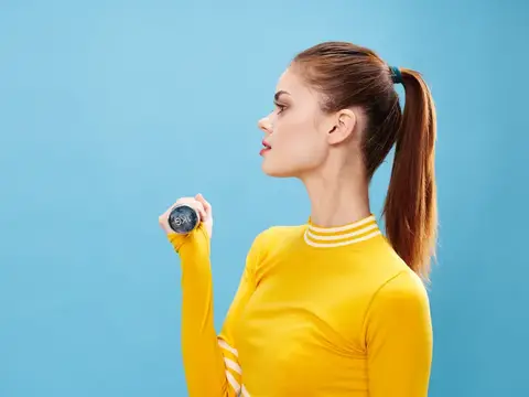 Молодая женщина с гантелью в желтой спортивной кофте на синем фоне