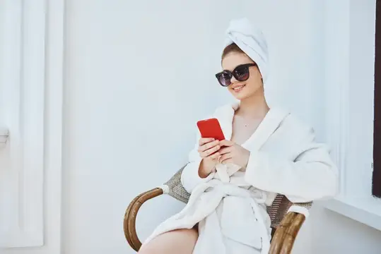 Улыбающаяся девушка в халате и солнечных очках смотрит в телефон