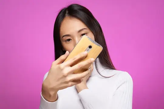 Азиатская девушка выглядывает из-за смартфона на розовом фоне
