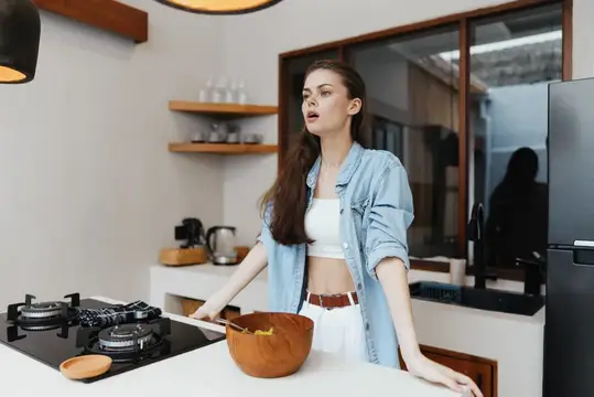 Молодая женщина в джинсовой куртке стоит на современной кухне и готовит полезную еду с помощью деревянной миски