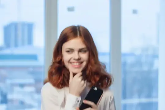 Рыжеволосая девушка улыбается, глядя на телефон у окна