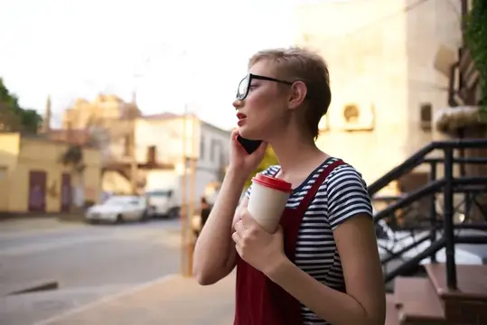 Женщина с короткими волосами, в очках, с чашкой кофе разговаривает по телефону