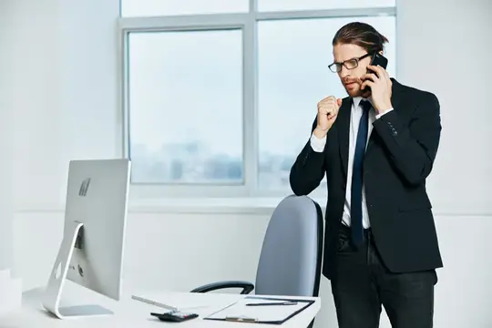 Мужчина в костюме радостно говорит по телефону у окна офиса