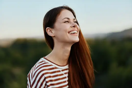 Улыбающаяся девушка с длинными каштановыми волосами на фоне зелёных холмов