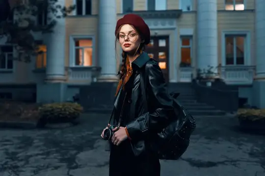 Молодая женщина в берете и кожаной куртке с фотокамерой на фоне старинного здания