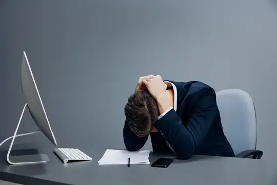 Красивый бизнесмен сталкивается с проблемами на работе, сидя за столом перед компьютером на сером фоне