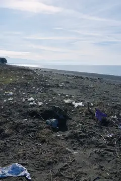 Пустынный скалистый берег с видимым мусором на берегу, море вдалеке и обломки, разбросанные по песку