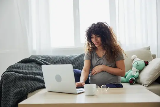 Беременная женщина с кудрявыми волосами работает за ноутбуком на диване