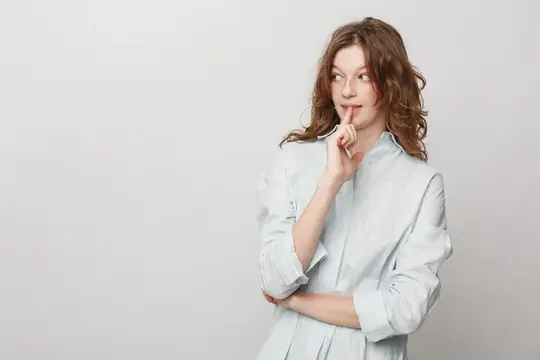 Любопытная женщина с вьющимися волосами в белой рубашке прикладывает палец к губам и смотрит в сторону