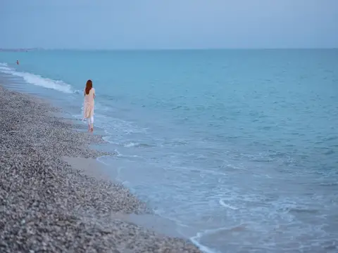 Девушка в светлом платье идёт по галечному пляжу у бирюзового моря