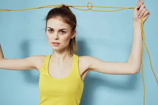 Спортивная женщина держит скакалку, на ней жёлтый топ без рукавов, она занимается гимнастикой на синем фоне