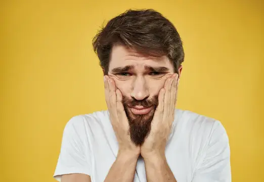 Уставший бородатый мужчина сжимает лицо руками на жёлтом фоне