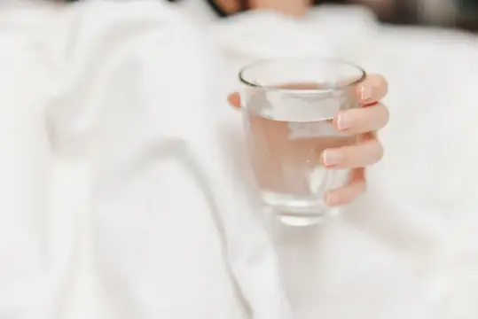Женская рука держит стакан воды на фоне белой постели