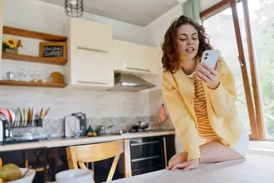 Кудрявая девушка в жёлтом кардигане смотрит в смартфон на кухне