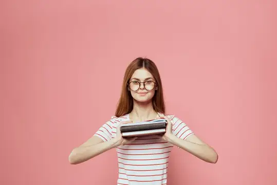 Рыжеволосая девушка в очках держит книгу на розовом фоне