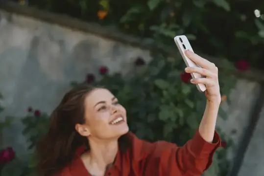Улыбающаяся темноволосая женщина делает селфи на фоне розовых кустов
