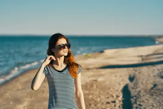 Рыжеволосая девушка в солнечных очках гуляет по пляжу у моря