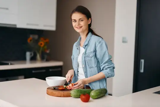 Улыбающаяся девушка в джинсовой рубашке режет овощи на кухне
