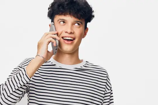 Молодой человек с радостной улыбкой разговаривает по смартфону, одетый в повседневную полосатую рубашку, с вьющимися волосами