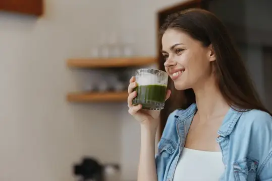 Улыбающаяся темноволосая девушка пьёт зелёный смузи на кухне