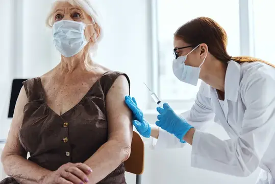 Медсестра в маске делает прививку пожилой женщине в светлом кабинете