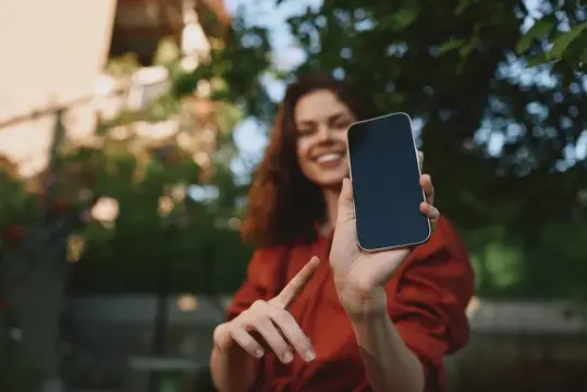 Улыбающаяся женщина в повседневной одежде делает селфи на смартфон на улице среди зелени и дневного света