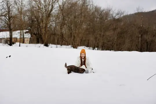 Рыжеволосая девушка в оранжевой шапке играет с собакой в снегу