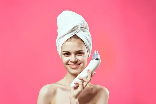 Улыбающаяся светловолосая девушка с полотенцем на голове держит косметический прибор на розовом фоне