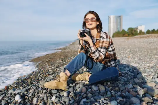 Улыбающаяся девушка с пленочным фотоаппаратом сидит на галечном берегу моря