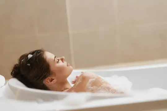 Безмятежная молодая женщина, погруженная в наполненную пеной ванну, излучает чувство покоя и расслабления в окружении современного дизайна ванной комнаты, дарящего тепло