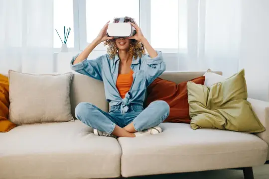 Улыбающаяся кудрявая женщина в VR-очках сидит на диване с подушками