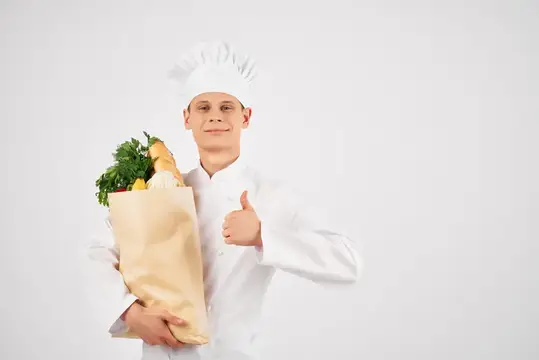 Молодой повар в белом колпаке с пакетом продуктов показывает большой палец вверх