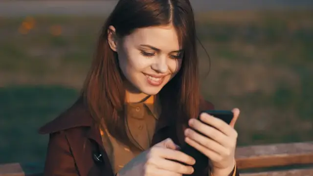Весёлая женщина в парке сидит на скамейке с телефоном в руках, общение