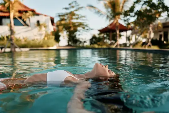 Портрет у открытого бассейна: женщина в купальнике дрейфует на воде, излучая отдых и беззаботную отпускную атмосферу на фоне тропических пальм и линии горизонта у моря
