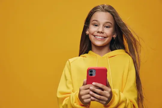Улыбающаяся девочка в жёлтом худи держит красный смартфон на жёлтом фоне