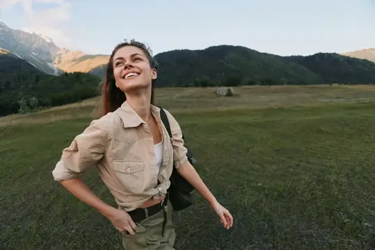 Женщина в светло-бежевой рубашке и оливковых шортах стоит на открытом лугу с небольшой сумкой на боку, заметно расслабленная и связанная с окружающей природой