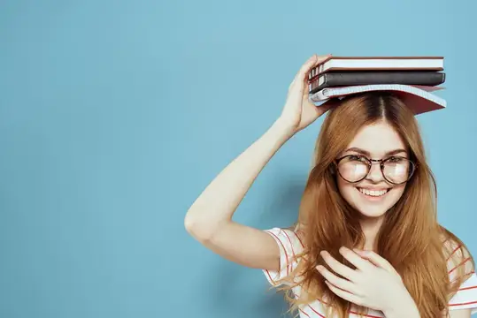 Рыжеволосая девушка в очках держит книги на голове и улыбается