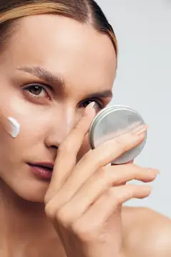Молодая женщина держит маленькое зеркальце, одновременно нанося увлажняющий крем на лицо, что подчеркивает личный уход, здоровье кожи и ежедневные бьюти-ритуалы в чистой обстановке
