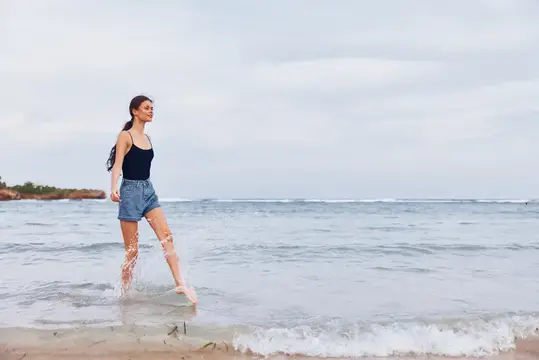 Молодая женщина босиком идёт по кромке воды в пасмурный день