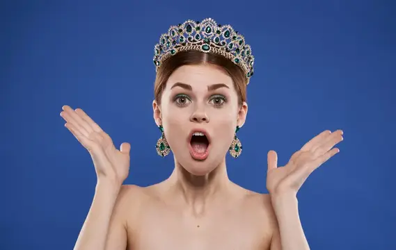 Красивая женщина в короне на голове, с макияжем, модель, делает жест рукой на синем фоне