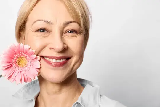 Улыбающаяся блондинка прижимает к щеке розовую герберу