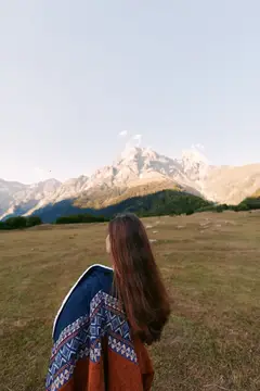 Женщина на лугу с пледом на плечах стоит лицом к горам на широком пейзаже