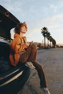 Женщина в стиле лайфстайл на городской дороге на закате: сидит на капоте автомобиля с вьющимися волосами