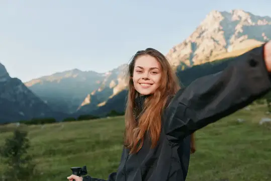 Фотограф наслаждается широким открытым пейзажем, улыбаясь на свежем воздухе, в то время как за ее спиной возвышаются скалистые пики, а солнечный свет освещает зеленые холмы и безмятежную долину