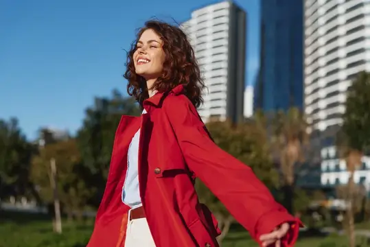 Молодая женщина с вьющимися волосами стоит в солнечном городском парке, раскинув руки