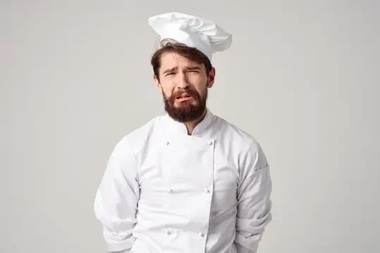 Усталый бородатый повар в белом колпаке и кителе