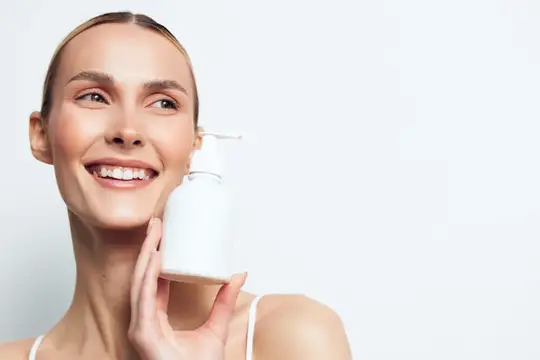 Улыбающаяся светловолосая женщина с белым флаконом косметики
