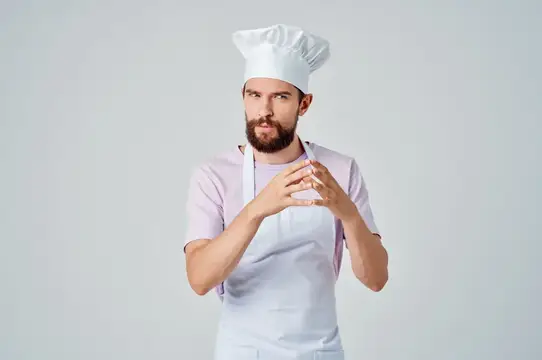 Бородатый мужчина-повар в колпаке и фартуке сложил руки на груди