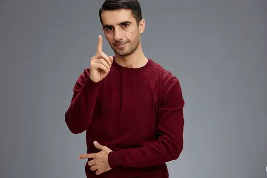 Успешный мужчина держит указательный палец у лица, жестикулируя руками, в красном свитере, на сером фоне